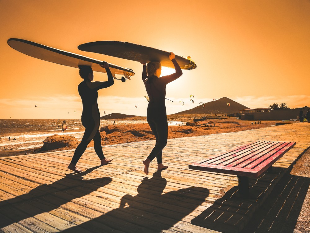 Surf girls go surfing. Women with surfboard on a beach at sunrise. Surfers and ocean. Silhouettes of two women walking along a beach at sunset. Sport image of longboard ina beautiful landscape