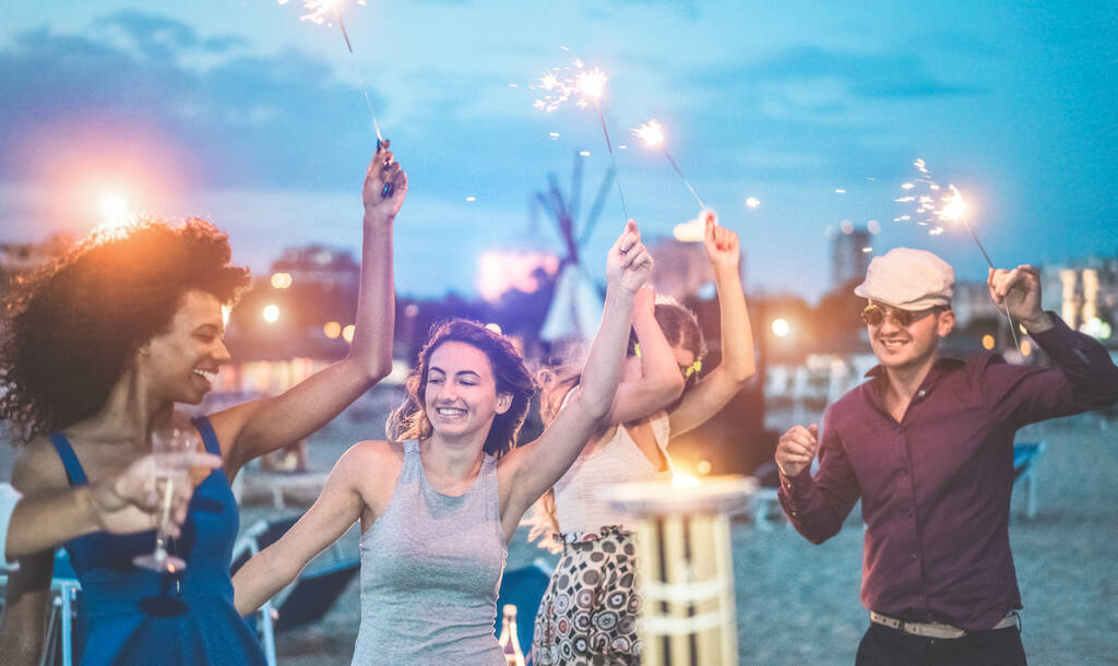 Happy friends making evening beach party outdoor with fireworks - Young people having fun dancing and drinking champagne - Soft focus on center woman hand - Vacation and nightlife concept